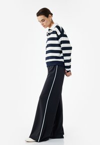 Mujer con pelo corto oscuro que lleva una camiseta de polo de rayas azul marino y blanca de manga larga, pantalones anchos negros con rayas blancas en los laterales y zapatos blancos.