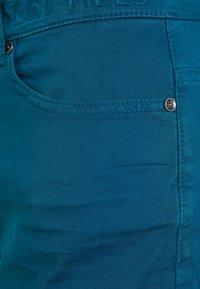 Tyrkysové jeansy z denimu s hladkou strukturou. Mají přední kapsu a kovový knoflík na pase. Diskrétní švy jako akcenty.