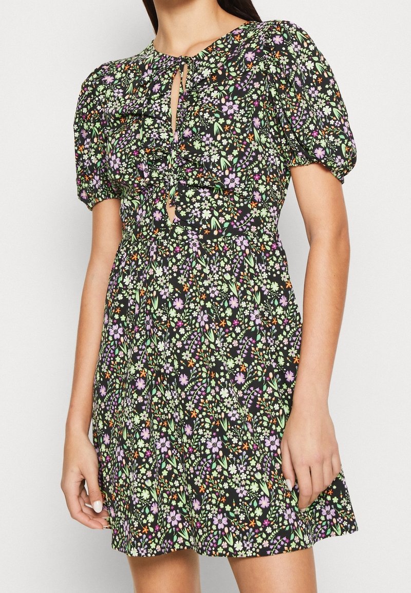 Femme portant une robe courte noire avec des manches bouffantes et un petit motif floral coloré, avec une ouverture en forme de clé à l'avant et une taille froncée.