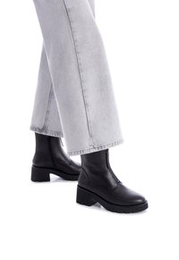 Botas negras de cuero hasta el tobillo con suela gruesa y cremallera lateral, combinadas con unos jeans de pierna ancha en gris claro. Las botas tienen una textura suave.