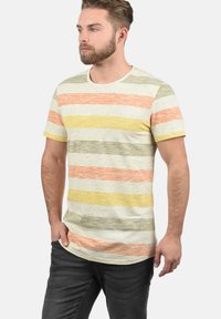 Juostuota t-shirt smėlio spalvos, su oranžinėmis, geltonomis ir žaliomis horizontaliosiomis linijomis. Trumpomis rankovėmis, apvaliu iškirpimu ir laisvu siluetu.