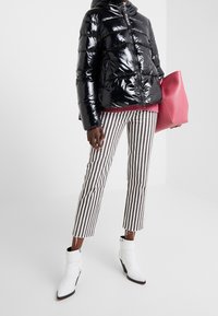 Czarna, błyszcząca kurtka puchowa, brązowe i białe pionowe paski na spodniach, białe botki za kostkę, trzymająca dużą różową torbę na tle białym.