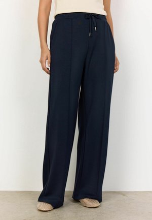 Femme portant un pantalon large bleu marine avec taille à cordon et des chaussures plates beige cloutées, debout sur un sol en béton.
