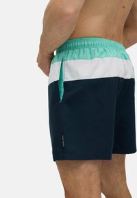 Bañador con un diseño mixto que combina secciones en color azul verdoso, blanco y navy. La cintura elástica incluye un cordón ajustable y bolsillos laterales.