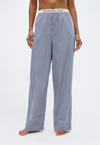 Άτομο με άνετο παντελόνι με κατακόρυφες λευκές και ναυτικές ρίγες, με κορδόνι μέσης με λογότυπο Polo Ralph Lauren, ξυπόλητο μπροστά σε απλό φόντο.