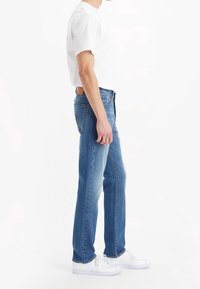 Jeans de mezclilla de lavado claro, corte recto, con un ligero desgaste y diseño clásico de cinco bolsillos, combinados con una camisa blanca de manga corta y zapatillas blancas.