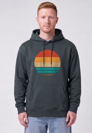 Young man with light skin and red hair wearing a dark hoodie with a vintage striped circle design in orange, yellow, cream, and green tones.