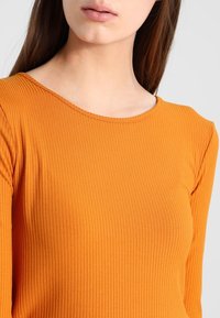 Eine Frau trägt ein langärmliges, geripptes oranges Oberteil mit einem runden Ausschnitt. Sie wird von den Schultern bis zu den Lippen vor einem schlichten Hintergrund gezeigt.