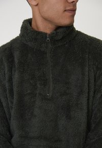 Man in een donkergroene zachte fleece pullover met een halve ritssluiting, zijwaarts gekeerd tegen een effen achtergrond.