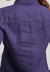 Person som bär en skrynklig lila långärmad skjorta, sedd bakifrån, med axellångt brunt hår synligt på ena sidan.