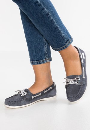 Pieds portant des chaussures bateau en daim bleu avec des lacets blancs, assorties à un jean denim bleu retroussé sur fond blanc.