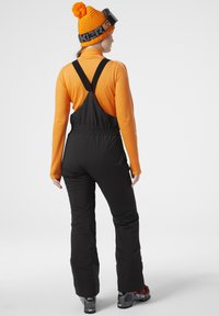 Overoles negros con tirantes ajustables, combinados con una camiseta de manga larga naranja y un gorro de lana a juego con un pompón.