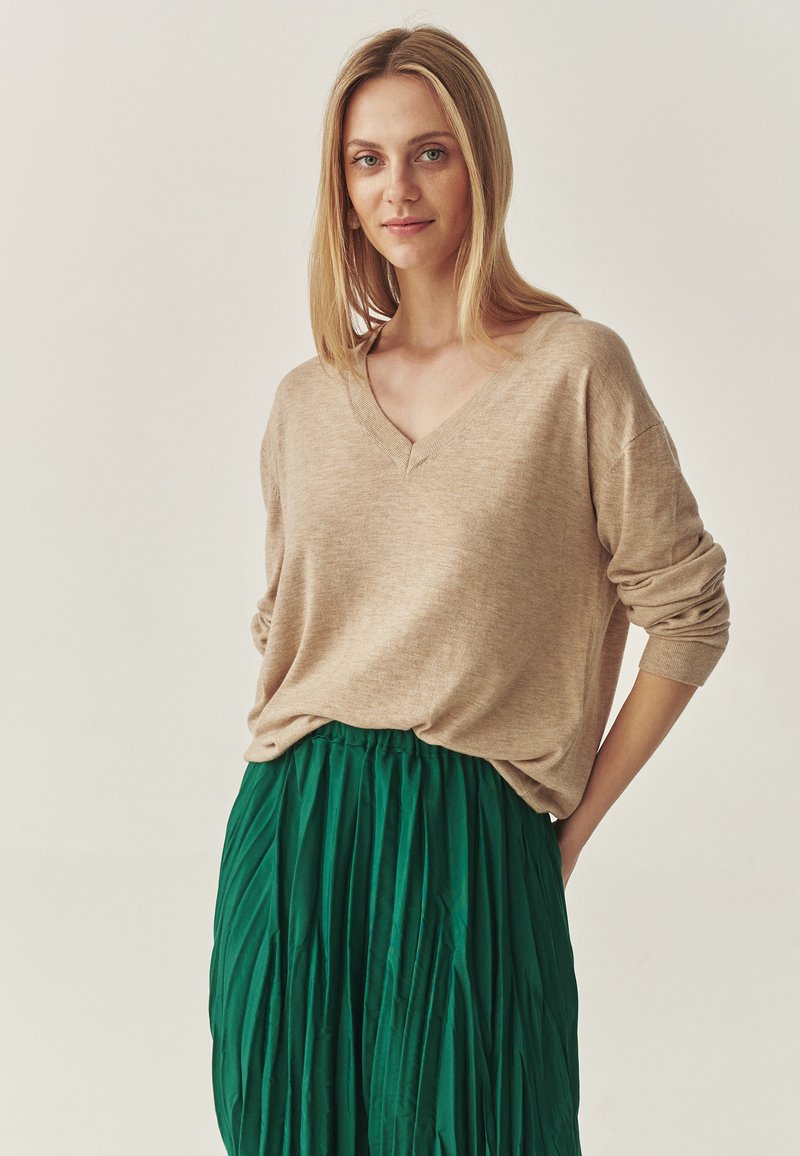 Beige gebreide trui met een v-hals en een los fitting, gecombineerd met een groene geplooide rok. De stof lijkt zacht en met een structuur te zijn.