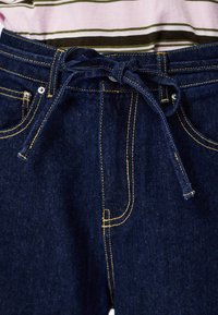 Vaqueros de mezclilla azul oscuro con costuras visibles en amarillo, bolsillos frontales y cintura con cordón ajustable. Tejido ligeramente texturizado con un diseño clásico.