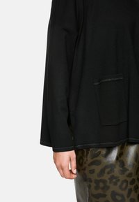 Schwarzes Oberteil mit lockerer Passform, das über eine Fronttasche und dekorative Nähte verfügt, kombiniert mit einem schimmernden Tiermuster-Rock.