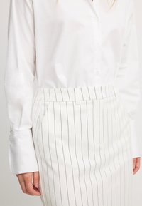 Chemise blanche à boutons avec des manches longues, associée à une jupe blanche à rayures avec des poches latérales et une taille ajustée.