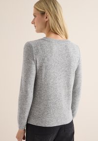Grå stickad tröja med långa ärmar och rundad halsringning. Mjuk textur med ett subtilt melerat mönster och åtsittande siluett.