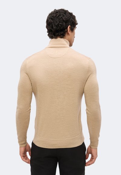 Homme vu de dos portant un pull à col roulé beige et un pantalon noir sur un fond clair.