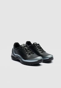 Paire de chaussures de sport noires avec des accents argentés, côtés perforés et lacets noirs, sur un fond gris clair uni.