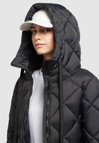 Jachetă puffer cu quiltă neagră, cu glugă mare și șnur. Închidere cu fermoar și strat interior alb contrastant vizibil dedesubt. Căciulă albă purtată.