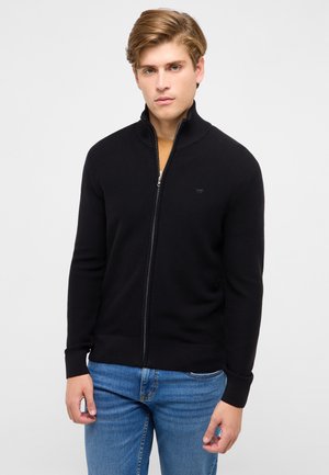 Junger Mann, der einen schwarzen Reißverschluss-Pullover über einem beigefarbenen Hemd und blauen Jeans trägt, vor einem schlichten hellen Hintergrund stehend.