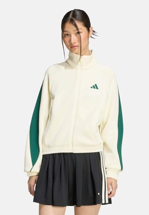 Femme portant une veste blanche zippée avec des rayures vertes sur les manches et une jupe plissée noire avec des rayures blanches sur les côtés, debout devant un fond uni.