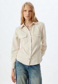 Camisa beige de manga larga, con botones a presión y dos bolsillos de solapa en el pecho. Tejido suave, diseño ligeramente ajustado y dobladillo curvado.