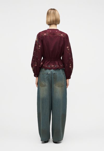 Γυναίκα με κοντά ξανθά μαλλιά φοράει μπορντό κεντημένο πουκάμισο και φαρδιά ξεβαμμένα μπλε τζιν, στέκεται μπροστά σε απλό λευκό φόντο.