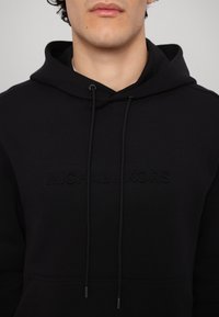 Hanorac negru dintr-un material moale, cu buzunar tip cangur, sfori de reglare și logo embossat. Textură netedă și croială lejeră.