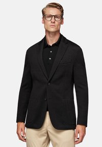 Black textured blazer with notch lapels, front buttons, and two front pockets, worn over a black polo shirt and beige trousers.