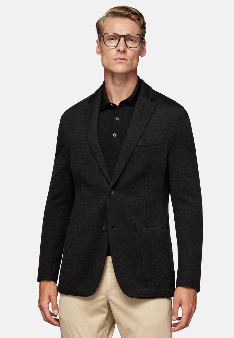 Black textured blazer with notch lapels, front buttons, and two front pockets, worn over a black polo shirt and beige trousers.