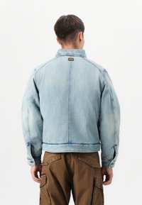 Veste en denim bleu clair avec un col, des poignets élastiques et une coupe droite. Le dos présente une couture centrale et un patch logo près du cou.