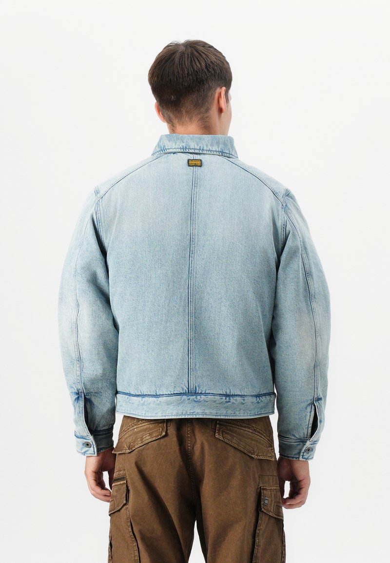 Lichtblauwe denimjack met een kraag, elastische manchetten en een rechte pasvorm. De achterkant heeft een centrale naad en een logopatch nabij de nek.