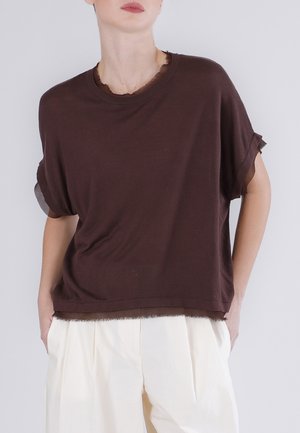 Personne portant un haut ample à manches courtes marron foncé avec des poignets retroussés et un pantalon plissé taille haute écru, mains dans les poches.