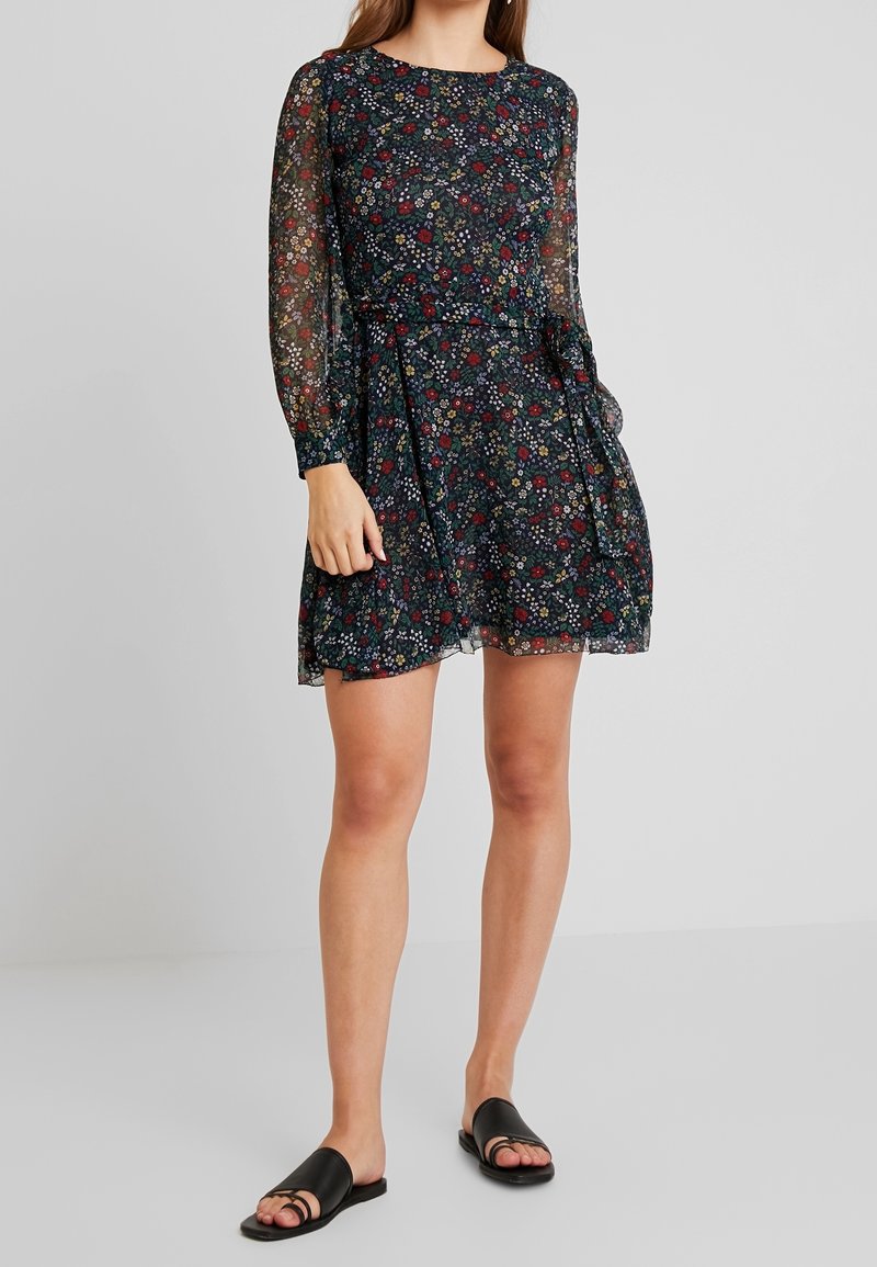 Femme portant une mini-robe à manches longues à motif floral sombre avec des manches transparentes et des sandales plates noires, debout devant un fond uni.