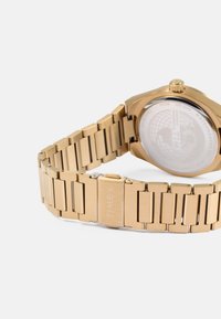 Relógio de metal dourado com pulseira ligada e caixa redonda. O relógio possui um fundo transparente com gráficos de um mapa mundi. Gravado "TIMEX."