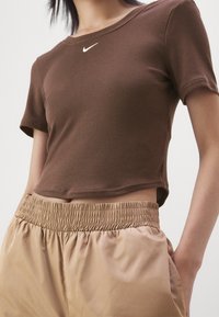 Person in einem maßgeschneiderten braunen Nike-Cropped-Shirt und beigen elastischen Hosen, steht mit einer Hand in der Tasche vor einem schlichten Hintergrund.