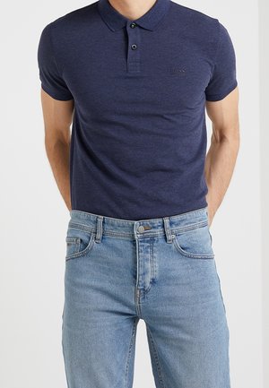 Homme portant un polo bleu foncé à manches courtes et un jean denim bleu clair, les mains reposant dans le dos.