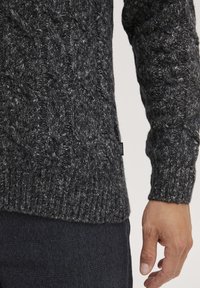 Tamsiai pilkas megztinis su pynimo raštu ir įdubimais ant rankovių, pagamintas iš tekstūruotos siūlės. Maža prekės etiketė matoma apačioje.
