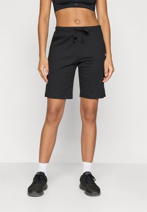Femme portant un short noir avec cordon, un soutien-gorge de sport noir, des chaussettes blanches et des baskets noires, debout devant un fond blanc uni.