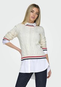 Balts jaukta adījuma džemperis ar sarkanu un tumši zilu svītru aprocēm, uzvilkts virs baltas krekla ar apkakli. Slim fit un teksturēts raksts. Tumši džinsi.