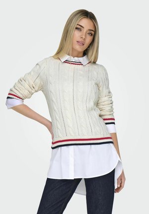 Weißer Kabelstrickpullover mit rot und marineblauen gestreiften Bündchen, über einem weißen Hemd mit Kragen getragen. Schmaler Schnitt und strukturiertes Muster. Dunkle Jeans.