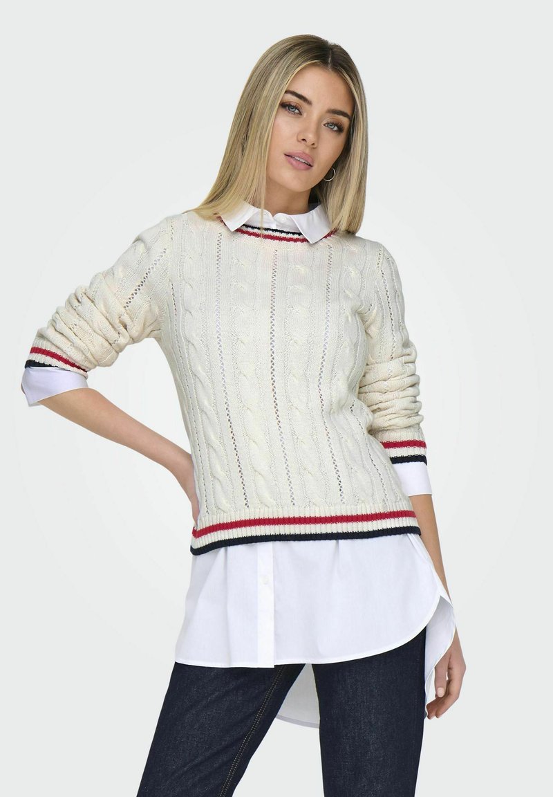 Balts jaukta adījuma džemperis ar sarkanu un tumši zilu svītru aprocēm, uzvilkts virs baltas krekla ar apkakli. Slim fit un teksturēts raksts. Tumši džinsi.
