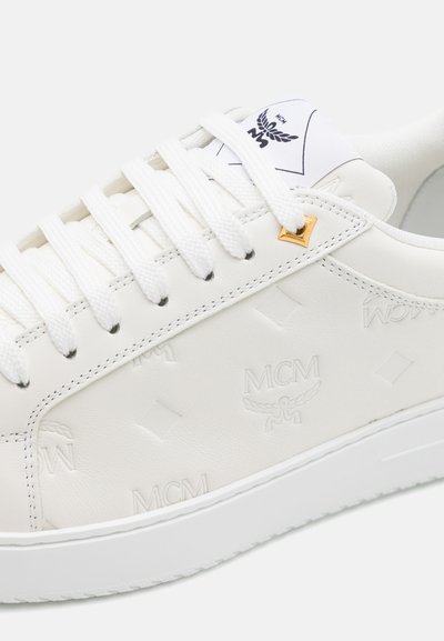 Zapatilla de cuero blanco con logotipo MCM en relieve y patrón de diamantes, que presenta cordones redondos blancos y un acento dorado cerca de los ojalillos.