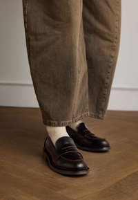 Leren loafers in donkerbruin met oranje stiksel. Gekomplementeerd met crèmekleurige sokken en loszittende beige broeken op een houten vloer.