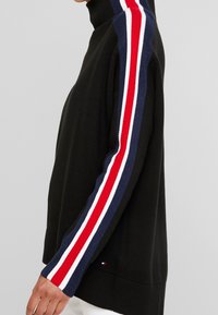 Černý rolák s námořnicky modrými, červenými a bílými pruhovanými akcenty podél rukávů. Pletená textura, volný střih, žebrované manžety a lem.