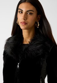 Czarna kamizelka z futerkiem sztucznym z wysokim kołnierzem, zapinana na zamek błyskawiczny, z siateczkowymi panelami po bokach. Modelka z dodatkiem złotych kolczyków-kołeczków.