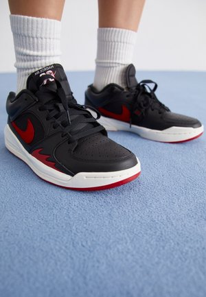 Zapatillas deportivas negras con acentos rojos, suela blanca, parte superior texturizada y área del dedo perforada; combinadas con calcetines blancos de caña corta.