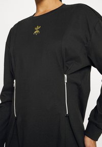 Svart sweatshirt med strukturerad yta, försedd med en liten grön logotyp och två fickor med dragkedja på framsidan. Långa ärmar med mudd.