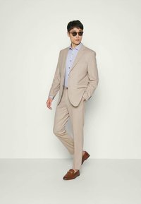 Man wearing beige suit, light blue shirt, brown loafers, and sunglasses, standing with one hand in pocket against plain background.
